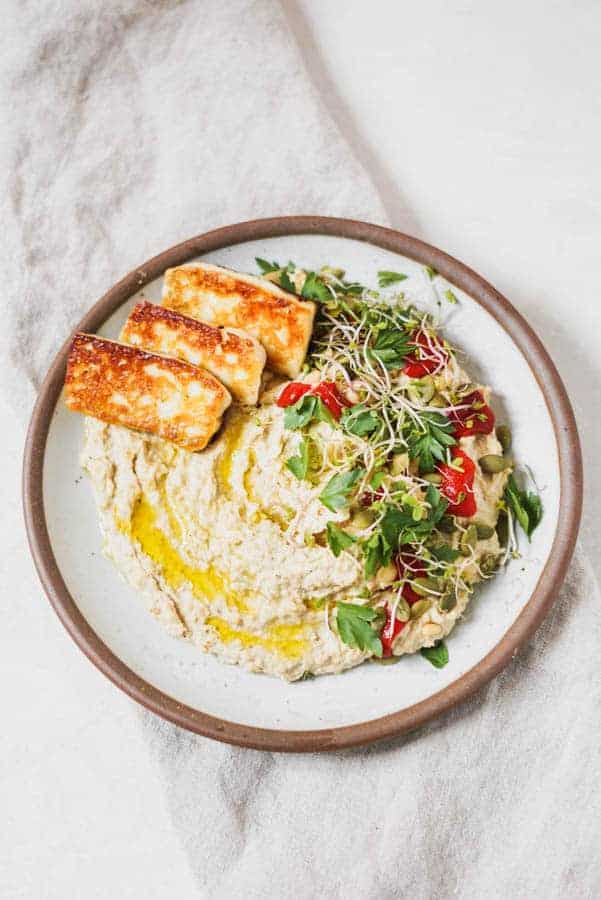 Brown plate full of hummus with herbs and cheese on top.